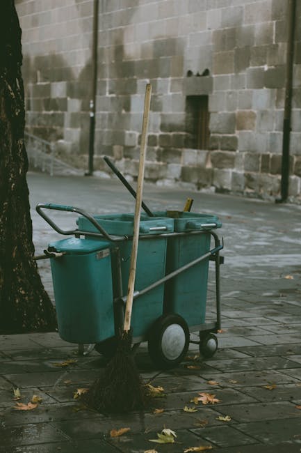 A street scene showing a blue cleaning cart with two large wheels, positioned on a paved sidewalk next to a tree trunk and a broom leaning against the cart. The cart contains cleaning supplies, and th