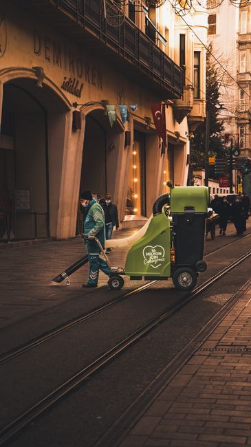 A street scene showing a blue cleaning cart with two large wheels, positioned on a paved sidewalk next to a tree trunk and a broom leaning against the cart. The cart contains cleaning supplies, and th