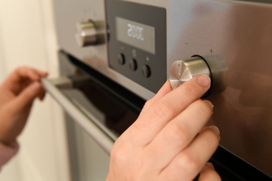 Close-up of a person's hand adjusting the temperature dial on a stainless steel oven in a domestic kitchen setting. The oven front features a digital display showing '20°C' and several control button